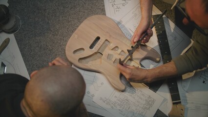 Top view of two male carpenters talking, making guitar according to drawings and taking...
