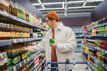 A young man in a casual white jacket and brown beanie analyzes a bottle of green soda in a...
