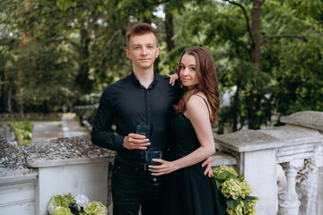 A stylish couple in black are standing on the balcony with a glass of wine in their hands against the background of green trees