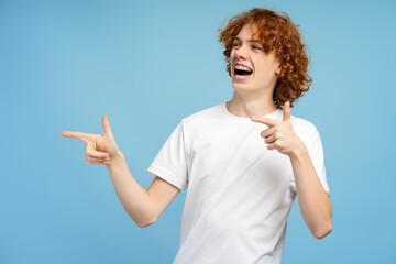 Smiling red haired curly boy wearing stylish casual t shirt pointing fingers on copy space