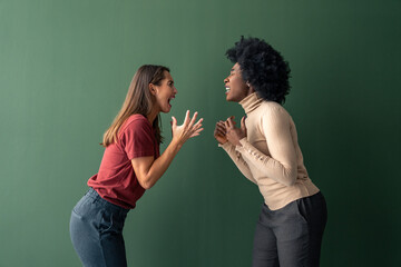 Two young interracial furious women yelling at each other against a green background in a studio.