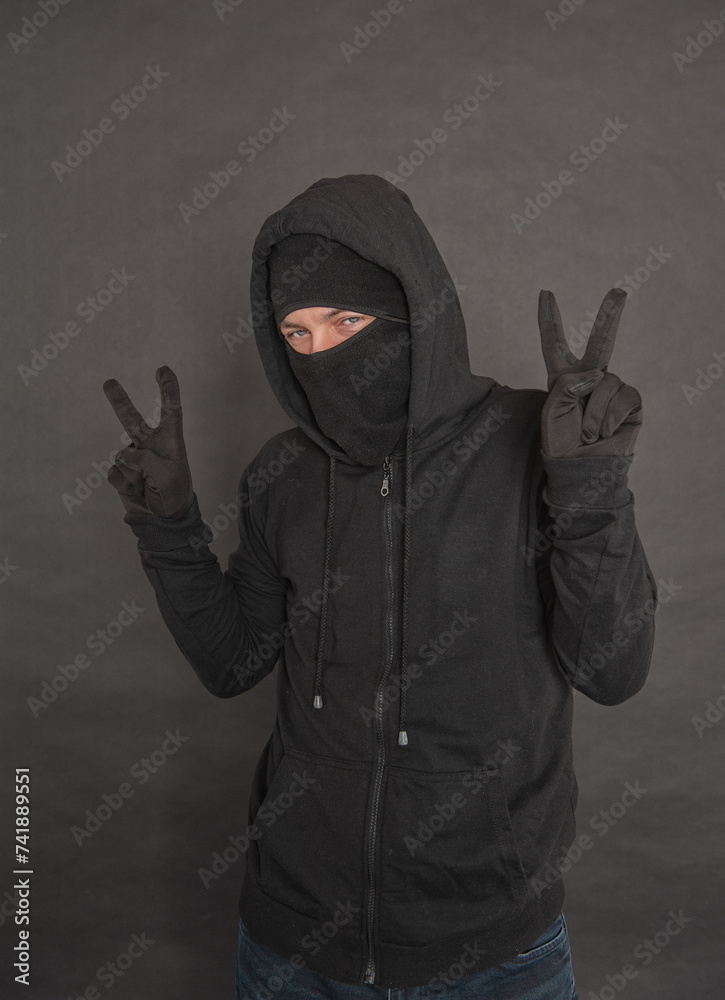 Wall mural Unrecognizable man in the black hoody with hood wearing balaclava mask showing two fingers victory gesture