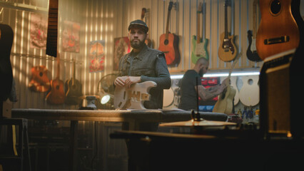 Male carpenter stands in modern woodworking workshop, holds wooden guitar body in hands, looks at camera. Colleagues make musical instruments in the background. Handcraft and small business. Portrait.