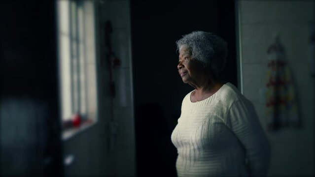 One contemplative elderly 80s woman standing by window in humble residence with thoughtful expression. Pensive senior lady with gray hair in quiet solitude