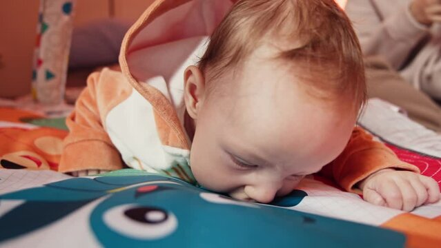 An infant in a cozy orange hooded onesie is intently exploring a colorful play mat with educational toys