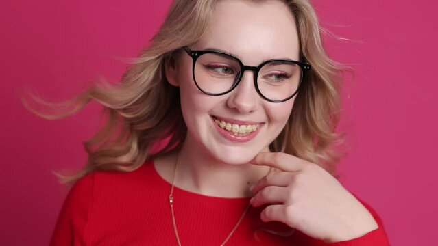 Cheerful young caucasian woman with transparent ceramic braces on teeth isolated on pink backdrop. Blond girl in eyeglasses looks at camera. Windy effect with fan. Beauty, health and fashion concept.