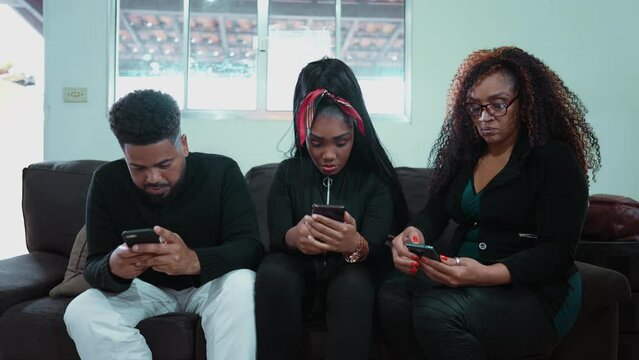 Three people staring at their phones seated on sofa indoors hypnotized by technology depicting social isolation and phone addiction. African American people texting, alone together