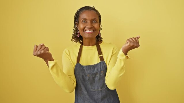 Triumphant middle age african american woman in apron, ecstatically screaming in confident victory, arms joyfully raised in celebration on a yellow isolated background