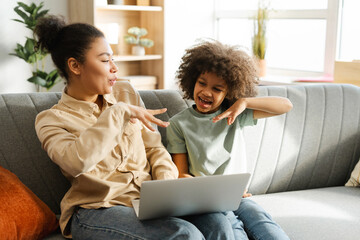 Smiling African American mother and her little daughter holding laptop, watching video together