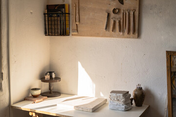 Warm sunlight bathes a tidy workshop corner, highlighting an array of ceramic tools, clay pieces, and a rustic pottery bowl on a white shelf