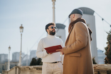 Two stylishly dressed businessmen having a discussion on a sunny day outdoors, one holding...