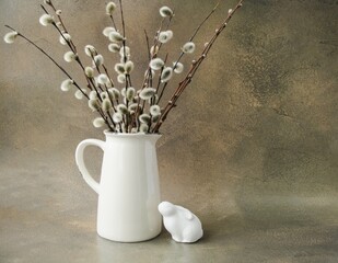 Easter composition on a brown concrete background on the table there is an Easter bunny with a willow twig in a vase.  Happy Easter holiday concept.  Space for copying text.  Front view.