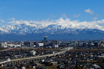 呉羽山から望む北アルプス立山連峰と富山市市街地駅を出る下り新幹線