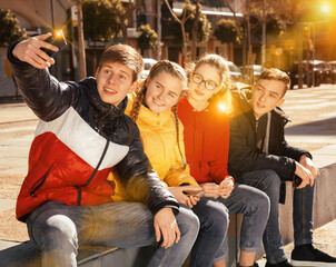 Fototapeta premium Portrait of friendly teenagers hanging out on streets of city on warm spring day, using phone to make selfie
