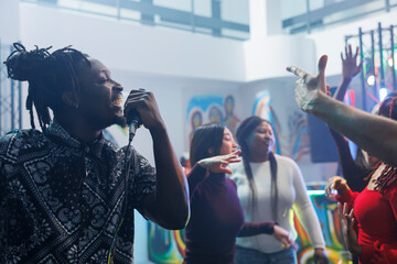 Smiling cheerful african american man singing in microphone while clubbing with friends. Young carefree clubber partying, holding mic and enjoying karaoke in crowded nightclub