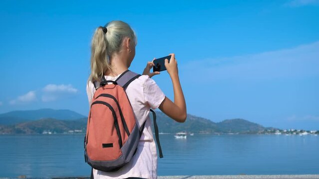 A young traveler photographs the sea. A teenage girl takes pictures of the blue sea on a smartphone.