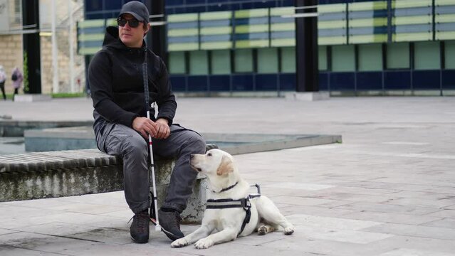 Blind man and his guide dog sitting on the street, handheld shot. Visually impaired people and assistance dogs concept.