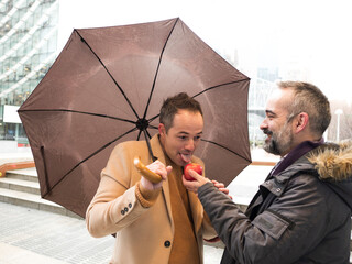 executive extends a red apple to a colleague who accepts it under an umbrella on a rainy day. The colleague begins to enjoy the apple, their shared shelter keeping them dry - 742005749