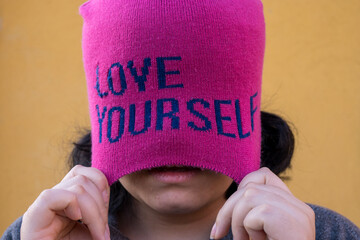 Girl wearing pink cap with self love message and pulling it, yellow background