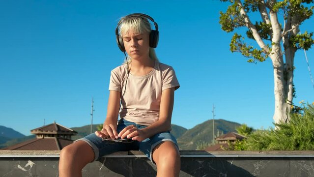 Worried blue haired girl in headphones. A view of pensive teen in headphones sitting in loneliness on the bay.