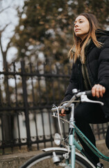A thoughtful young female in warm clothing stands with her bicycle in a city park, exuding a sense of solitude and reflection amid autumnal scenery.