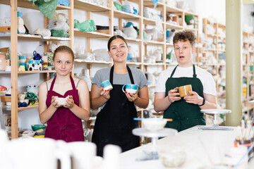 Young woman teacher and students teenagers girl and boy posing with ceramic cups in ceramic workshop