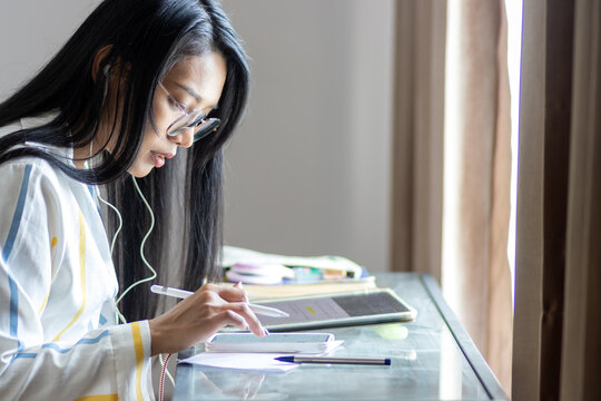 Teaching online - A young woman works with a tablet in a dark room lit by daylight from a window