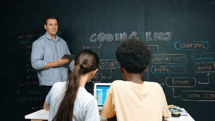 Caucasian teacher answering student question while holding tablet. Caucasian girl pointing at blackboard with code or prompt while laptop display system screen at STEM technology class. Edification.