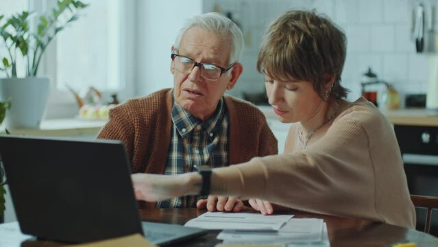 Elderly man learning how to pay bills online on laptop with assistance of young female social worker visiting him at home