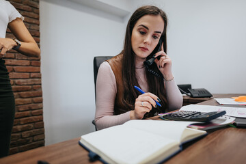 Focused professional woman on phone call in office environment with colleague in background.