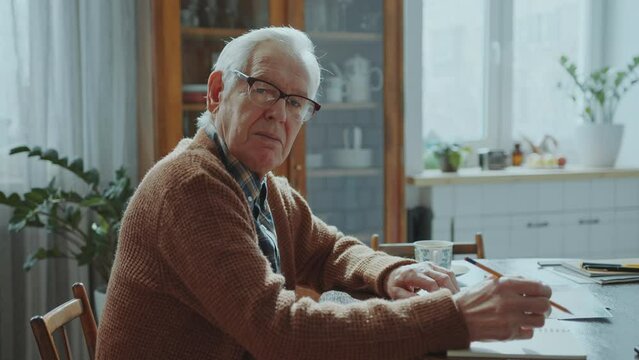 Elderly gray-haired man in glasses sitting at table with paper bills and copybook, holding pencil and looking at camera during budget planning at home. Video portrait