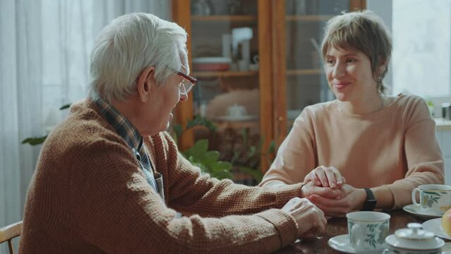 Young caring woman holding hands of senior granddad and talking to him, giving emotional support during visit at home