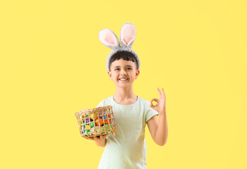 Cute little boy wearing bunny ears with basket of Easter eggs showing OK on yellow background