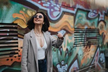 A fashionable young girl wearing sunglasses smiles as she walks by a colorful graffiti wall on a city street during fall.