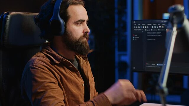 Smiling developer listening music while doing programming during remote job shift in home office. Upbeat IT admin enjoying audio podcast, working on PC in apartment late at night, close up