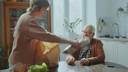 Female social worker taking off groceries from paper bag on kitchen table and speaking with senior man when helping him with daily living at home