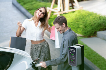 Young couple travel with EV electric car charging in green sustainable city outdoor garden in...