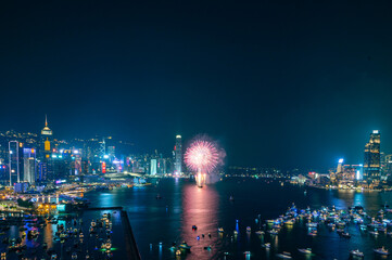 Fototapeta na wymiar Fireworks burst brightly against the night sky above a city skyline, reflecting in the calm water below