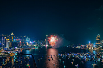 Fireworks burst brightly against the night sky above a city skyline, reflecting in the calm water...