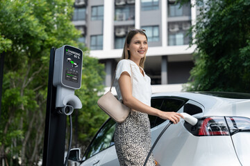 Young woman travel with EV electric car charging in green sustainable city outdoor garden in...