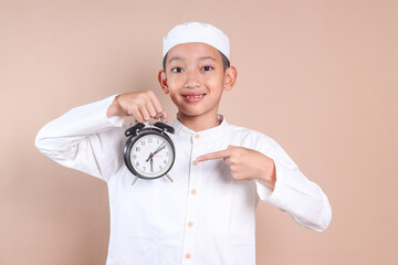 Cheerful Muslim boy wear skullcap, pointing at alarm clock that showing iftar time