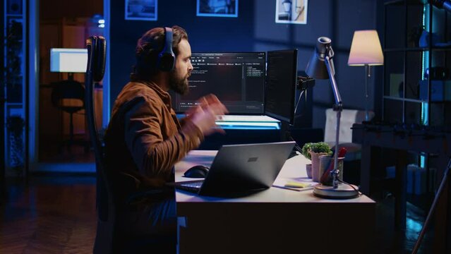 Technician focused on typing complex code in neon lit home office, developing software application. It specialist removing headphones to better concentrate on difficult task, camera B