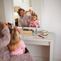 Fototapeta premium Mom, daughter and playing with hair in mirror for beauty, care and helping or getting ready. Happy mother, family and girl or kid with rollers for hairstyle, fashion and bonding together at home