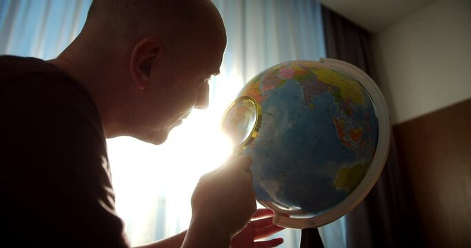 Man looks through a magnifying glass at a globe. Man using globe sits at home looking for right country on globe. 4K