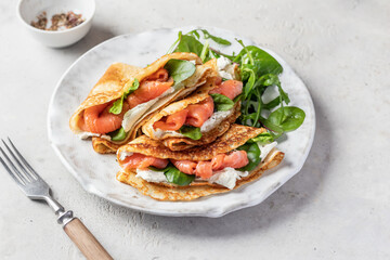 Crepes or thin pancakes with smoked salmon, soft cheese and spinach on a plate on gray textured...