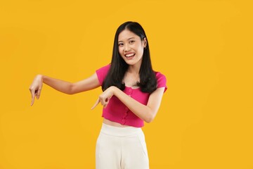 Obraz premium Photo of a cheerful young Asian woman wearing a casual pink t-shirt, using her finger to point down or empty copy space while standing. Isolated on a yellow background. Advertise concept.