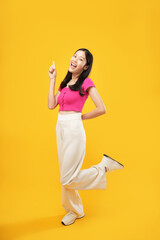 Full-length portrait of amazed excited young Asian woman 20s in a pink casual t-shirt pointing index fingers aside up on mock-up copy space isolated on Yellow color wall background studio