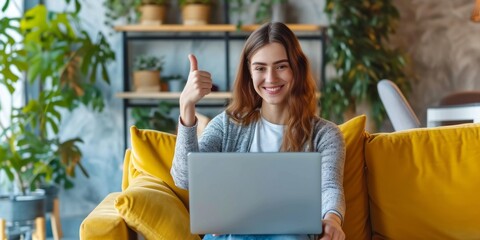 A young girl with a smile is working on her laptop. Alone woman sitting on a yellow sofa. Woman showing thumbs up.
