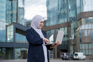 Fototapeta premium Thoughtful business woman in hijab and suit is holding a laptop outdoors.