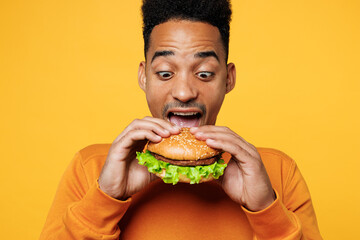 Close up young surprised man wear orange sweatshirt casual clothes open mouth eat bite burger...